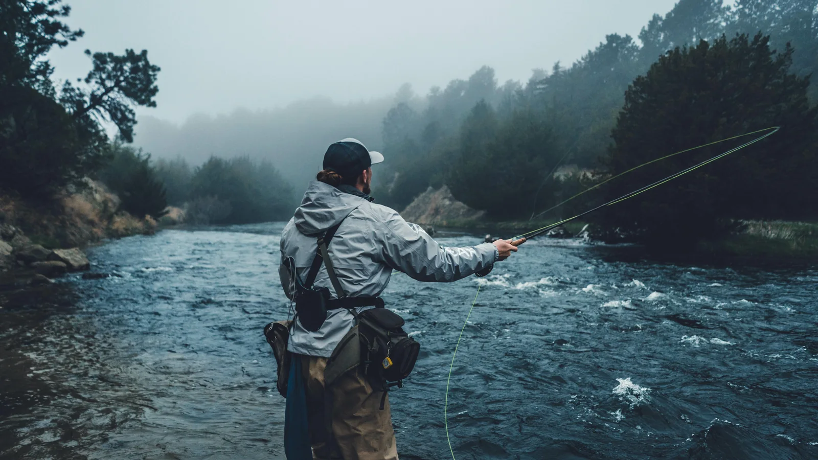 Un pêcheur à la mouche, de dos, lance sa ligne dans un courant de rivière par un matin de brume.
